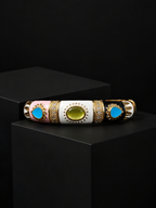 A decorative cylindrical bracelet or bangle displayed horizontally on a black pedestal. The bracelet has three main color sections: light pink, white, and black, separated by ornate gold filigree borders.