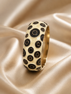 A wide gold band ring decorated with a pattern of black and cream concentric circles, resting on draped champagne-colored satin fabric.