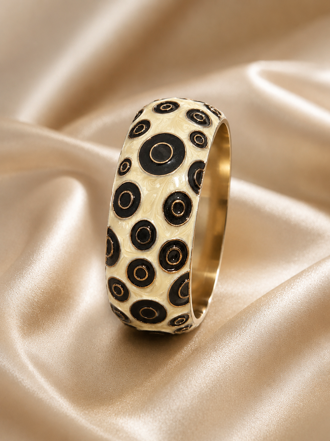 A wide gold band ring decorated with a pattern of black and cream concentric circles, resting on draped champagne-colored satin fabric.