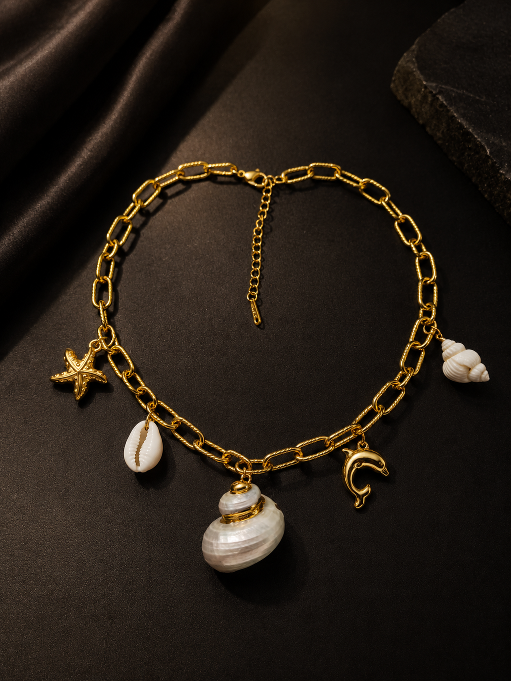 A gold chain necklace with shell and sea-themed charms, including a starfish, a spiral shell, a crescent moon, and a larger shell pendant, displayed on a dark textured background.