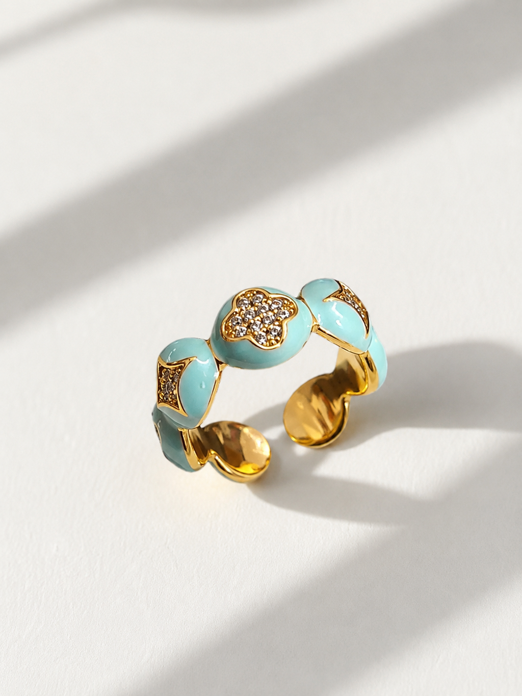 A gold-plated open cuff ring decorated with light blue enamel segments and small clear rhinestones, photographed on a white surface with soft diagonal shadows.