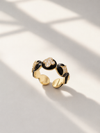 A gold and black enamel open ring with floral details, photographed on a light surface with soft window shadows.