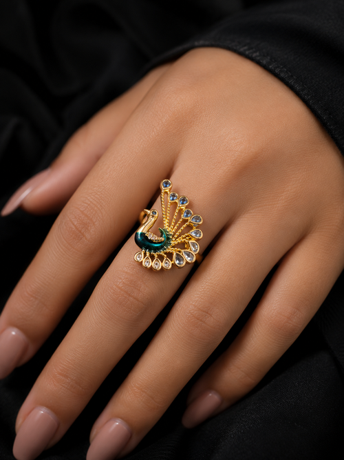 A gold-toned ring designed like a peacock with a green body and blue and clear gemstone details, shown worn on a hand against a black fabric background.