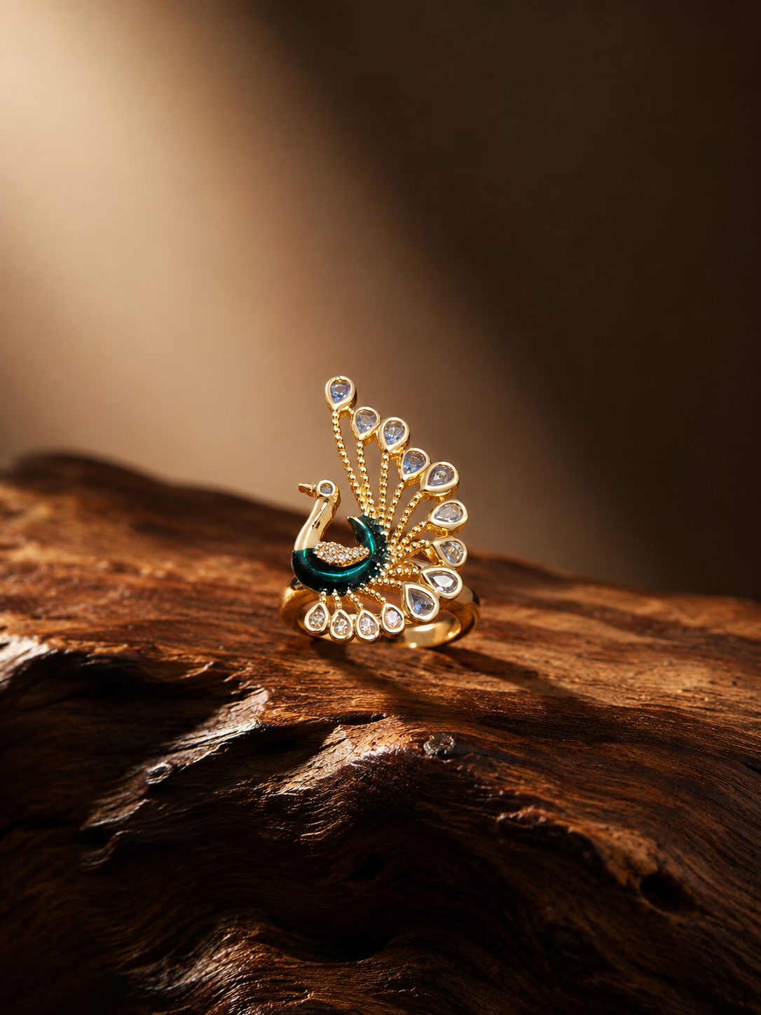 A gold-toned peacock ring with a dark teal enamel body and clear gemstones in the feather tips, displayed on a textured piece of dark wood against a warm, softly lit brown background.