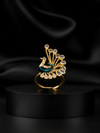 A gold-toned peacock ring with a teal enamel body and clear gemstone accents, displayed on a black cylindrical pedestal against a black satin background.