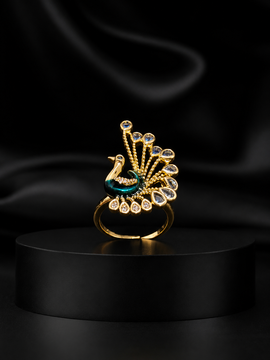A gold-toned peacock ring with a teal enamel body and clear gemstone accents, displayed on a black cylindrical pedestal against a black satin background.