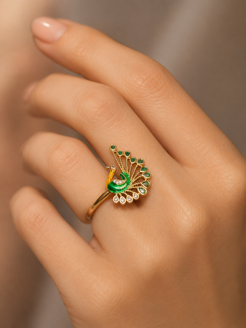 A gold ring designed like a peacock with green enamel and green gemstone accents, worn on a person's finger.