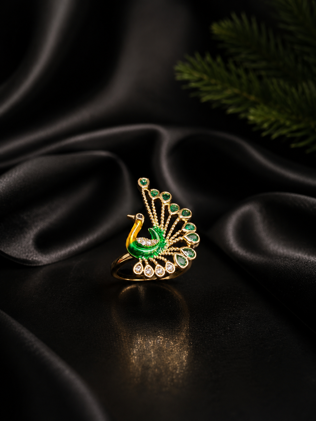 A gold ring designed as a peacock with a green and gold body and a fanned tail of green stones, displayed on a draped black satin surface with a dark green pine branch in the background.