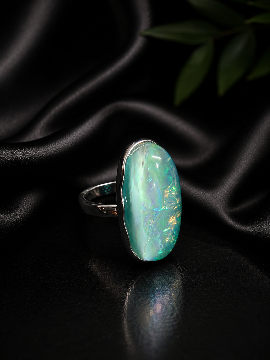 A silver ring featuring a large oval cabochon opal gemstone with iridescent green, blue, and gold flashes, displayed on a draped black satin fabric background.