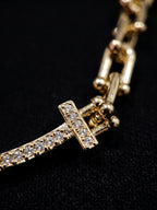 Close-up of a gold-tone chain bracelet featuring a curved bar set with small clear stones and a T-shaped clasp, photographed against a black background.