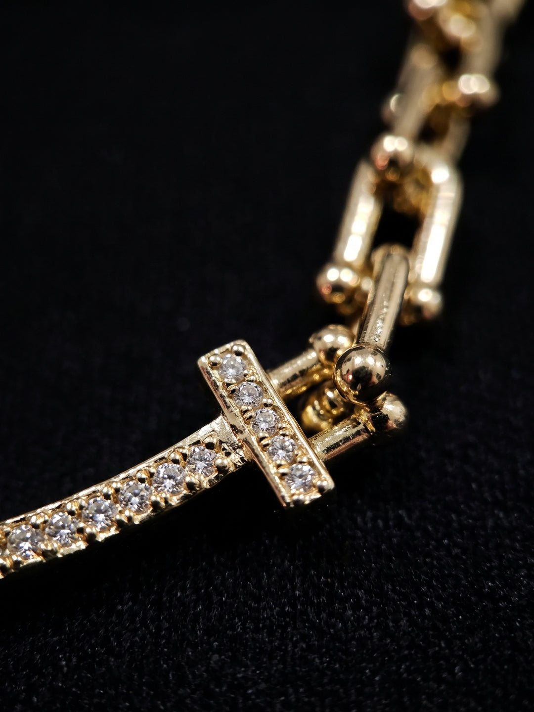 Close-up of a gold-tone chain bracelet featuring a curved bar set with small clear stones and a T-shaped clasp, photographed against a black background.