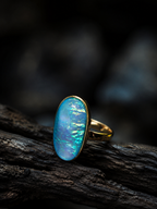 A gold ring with an oval blue iridescent opal stone, resting on a dark textured piece of wood against a dark, blurred background.