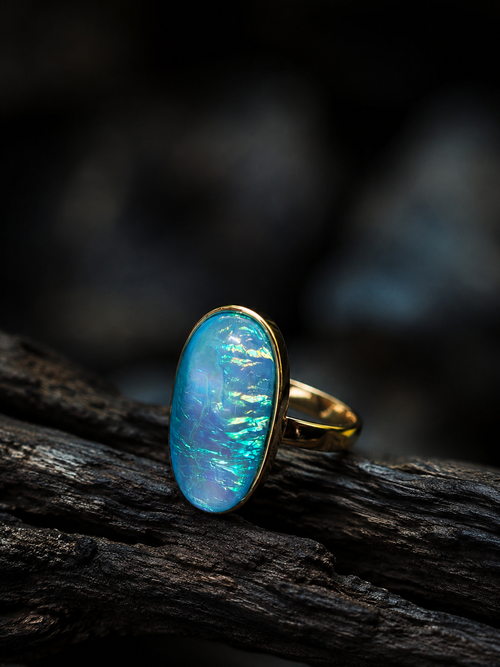 A gold ring with an oval blue iridescent opal stone, resting on a dark textured piece of wood against a dark, blurred background.