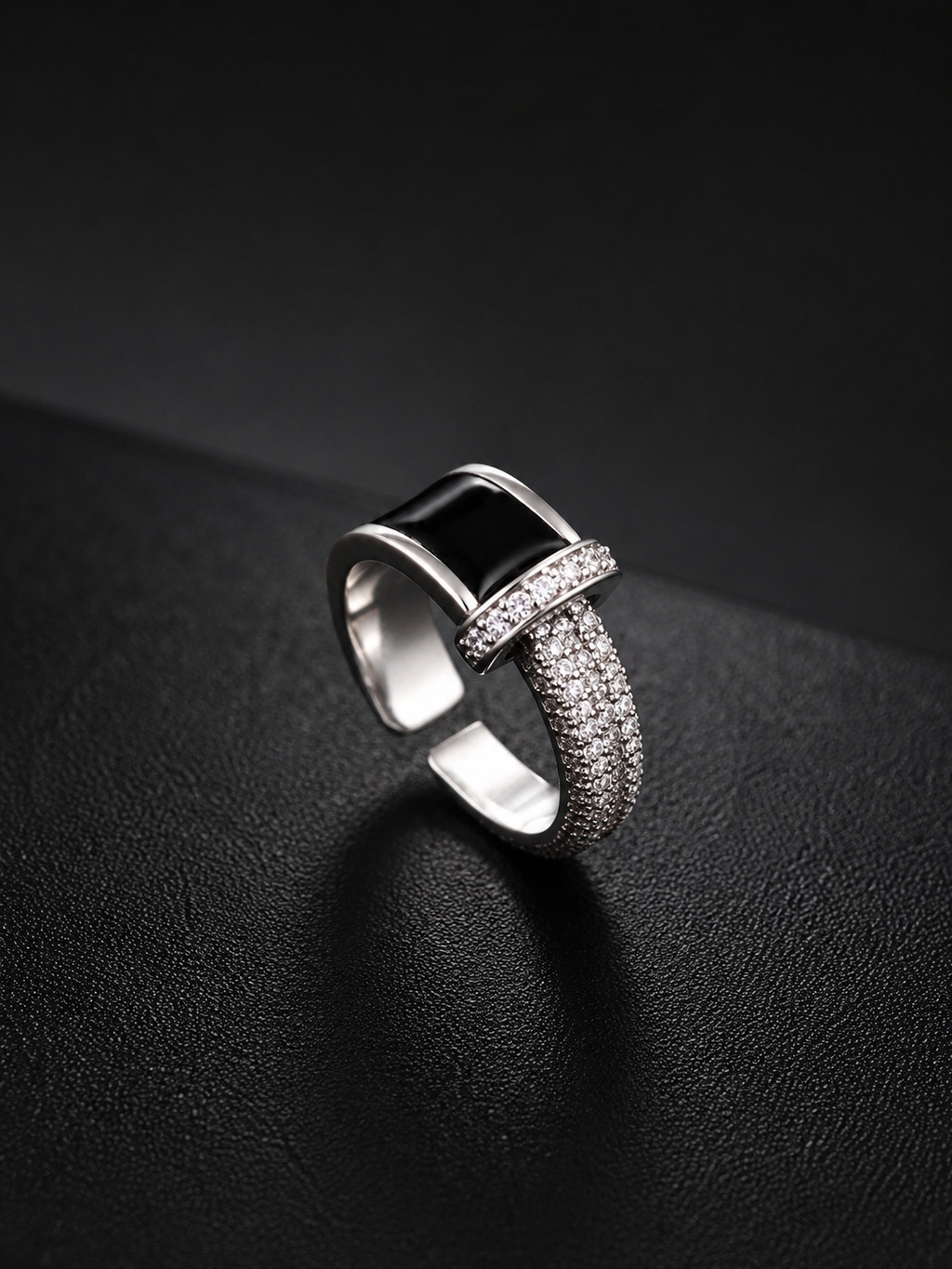 A silver-toned geometric ring with a rectangular black inlay and pavé-set clear stones, photographed on a dark textured surface.