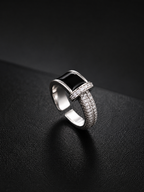 A silver-toned geometric ring with a rectangular black inlay and pavé-set clear stones, photographed on a dark textured surface.
