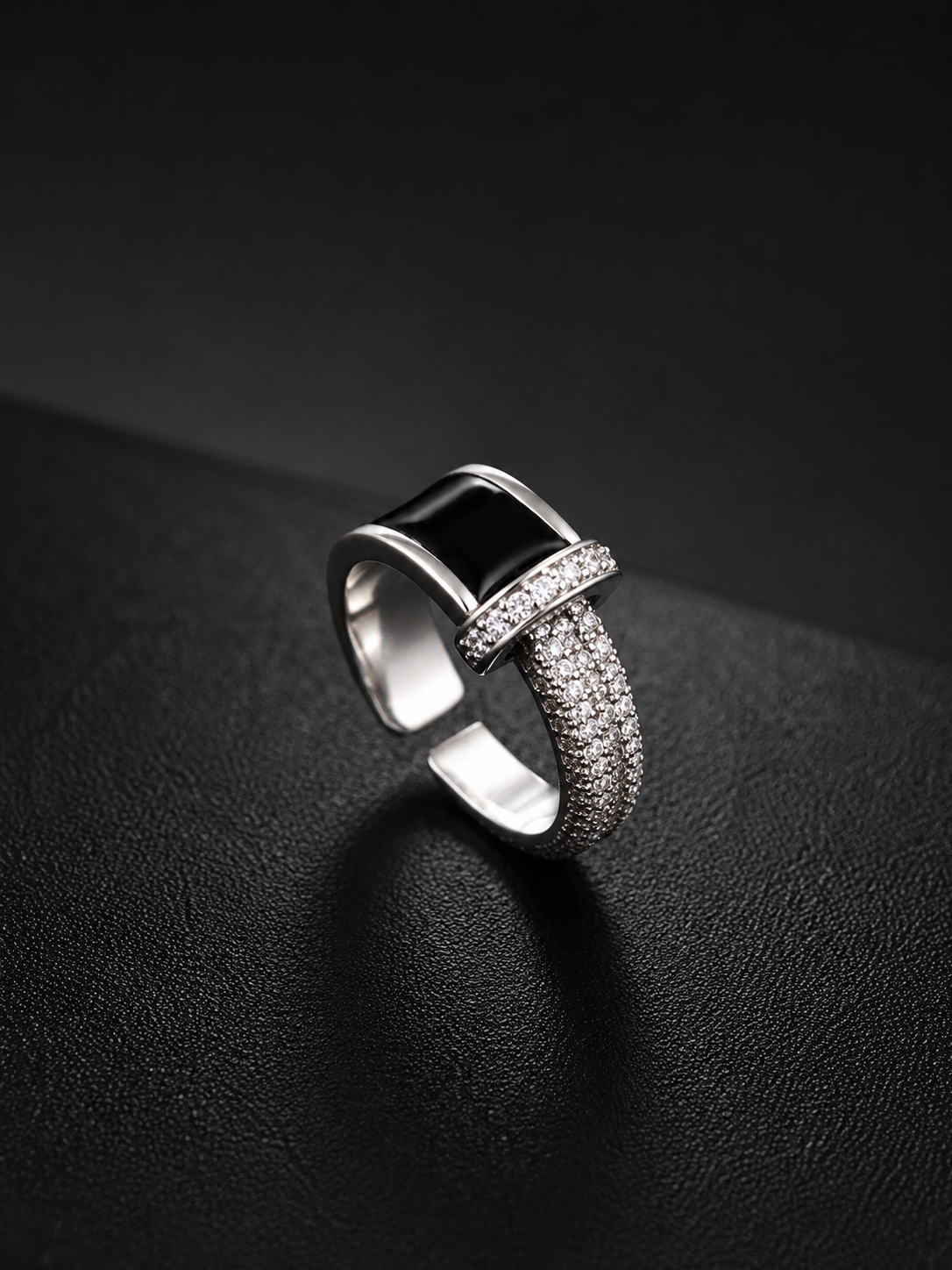 A silver-toned geometric ring with a rectangular black inlay and pavé-set clear stones, photographed on a dark textured surface.