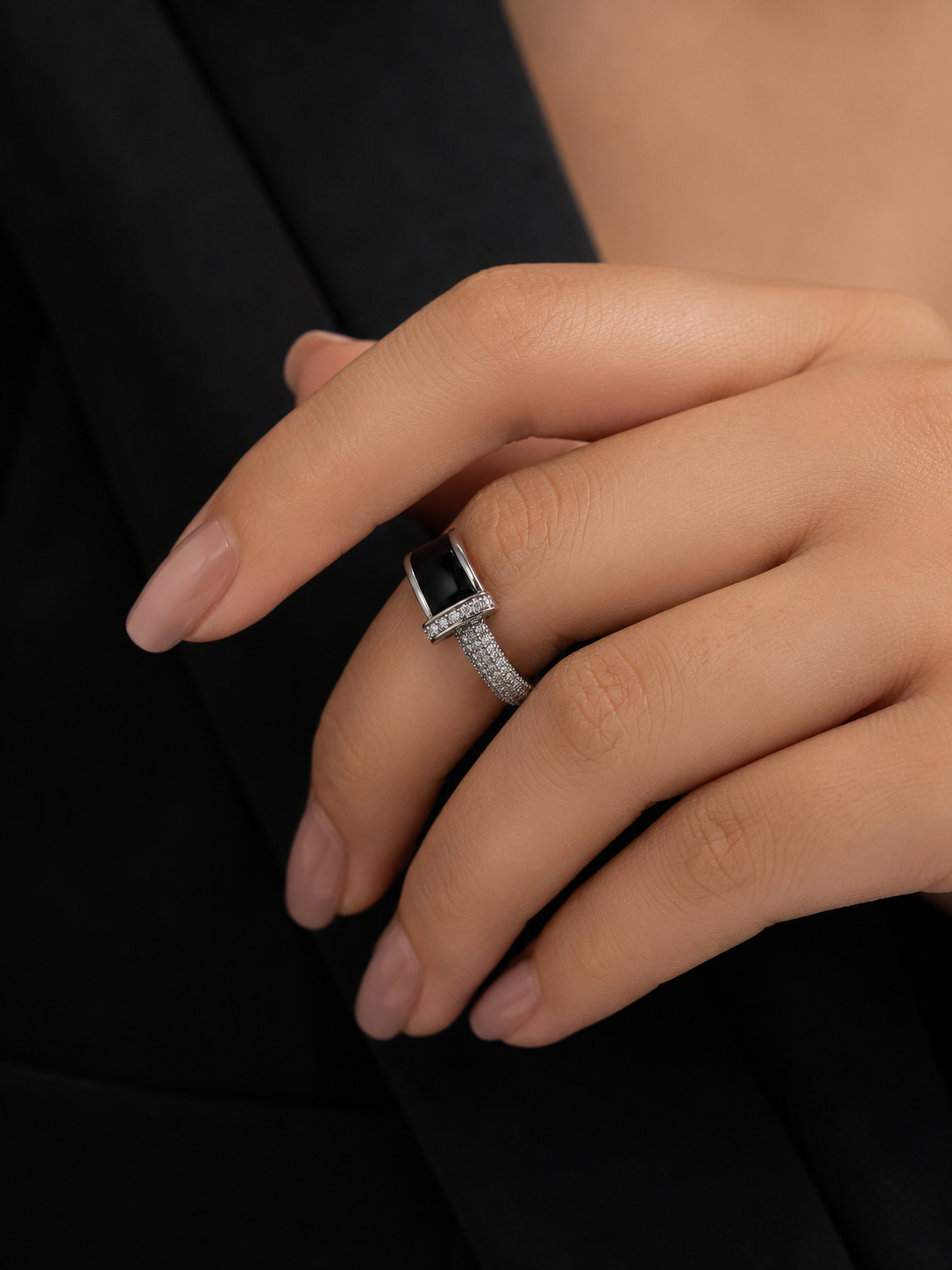 A close-up of a hand wearing a silver ring with a rectangular black inlay and small pavé-set stones, against a dark black blazer background.