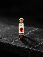 A highly detailed, vintage-style cylindrical ring or decorative piece featuring white enamel, pink enamel bands, black enamel sections, gold-toned metal borders, and red and blue gemstones, standing upright on a dark textured stone slab against a black background.