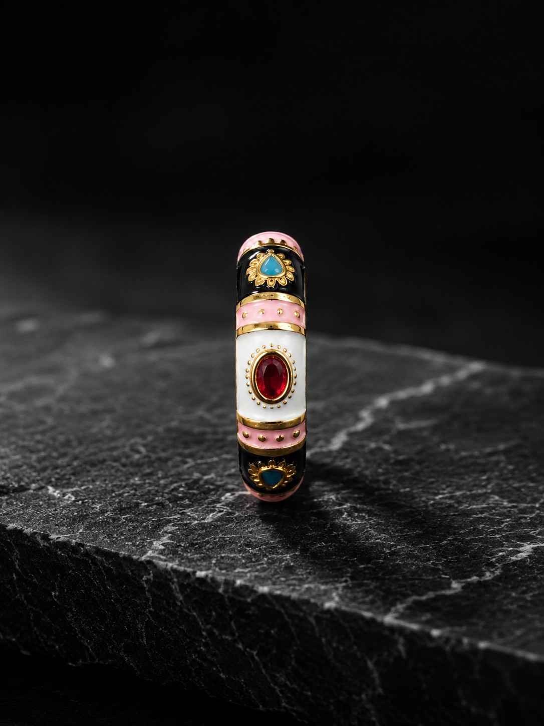 A highly detailed, vintage-style cylindrical ring or decorative piece featuring white enamel, pink enamel bands, black enamel sections, gold-toned metal borders, and red and blue gemstones, standing upright on a dark textured stone slab against a black background.