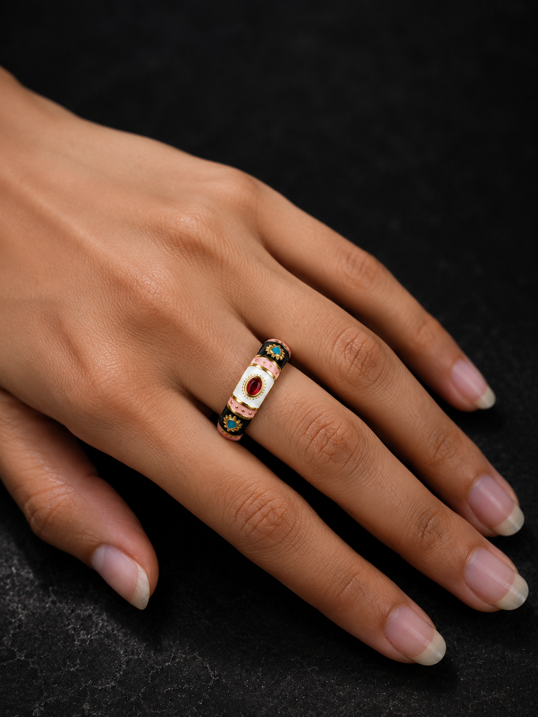 A close-up of a hand wearing a gold ring with a central white stone and small green and red accents on a dark textured background.
