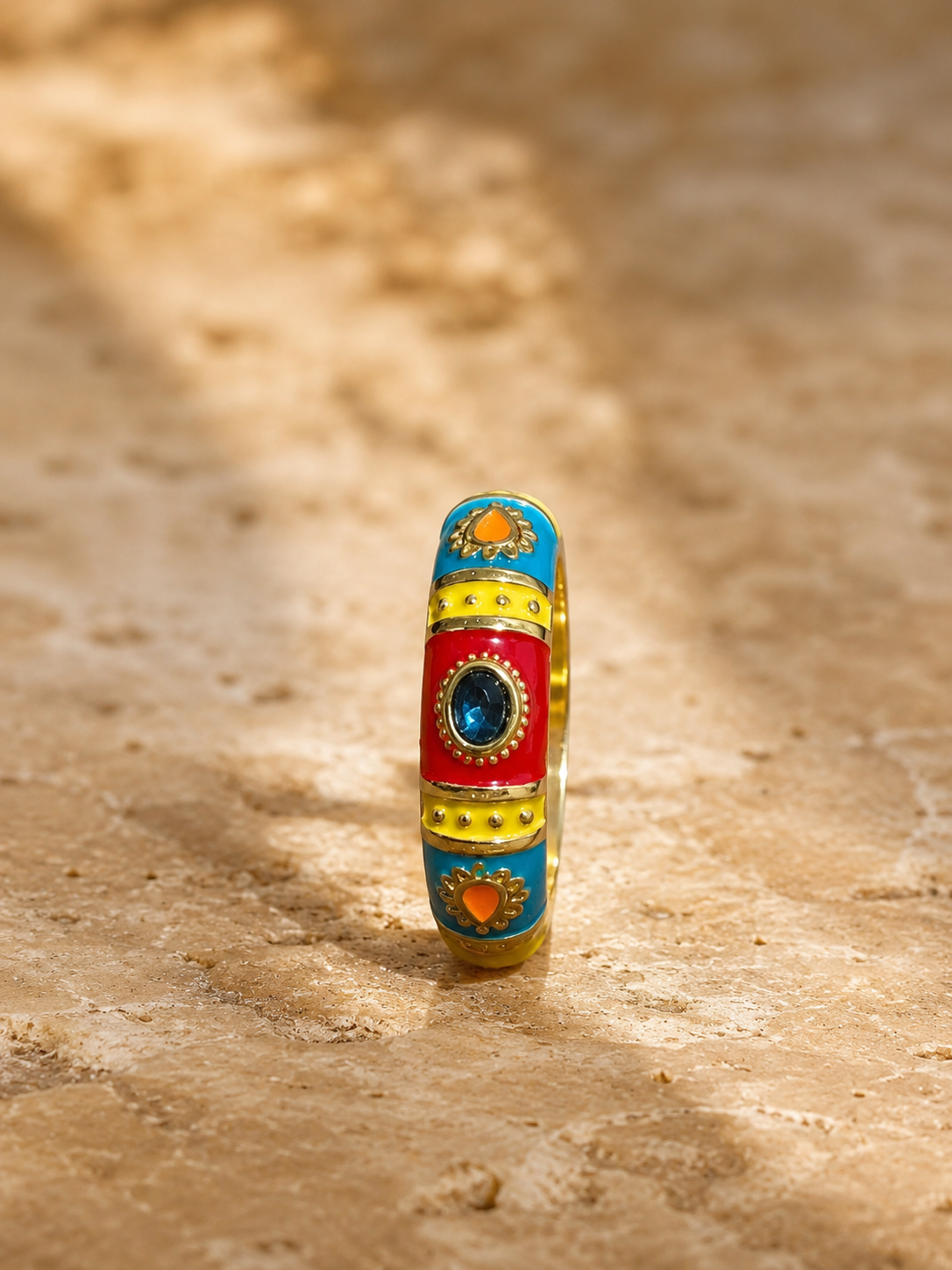 A colorful cylindrical ring with gold-toned metal, red enamel, blue accents, and a central blue stone, standing upright on a sandy surface.