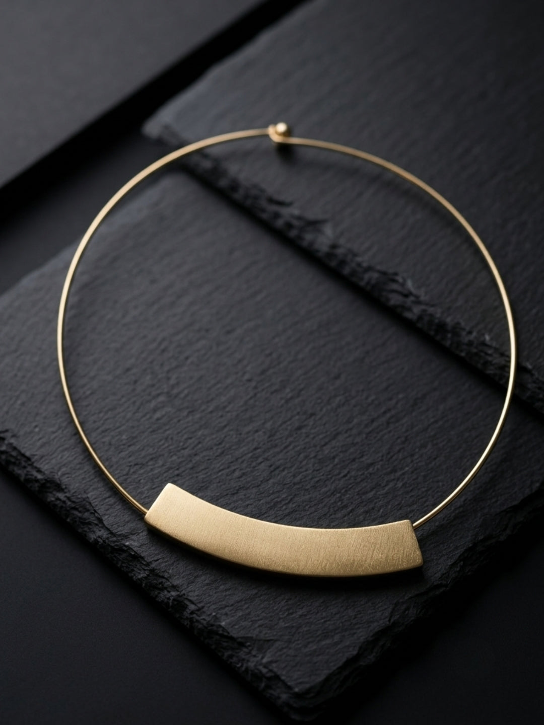A minimalist gold-toned necklace featuring a thin circular wire design and a brushed, curved horizontal bar pendant, displayed on a dark textured slate background.