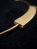 A close-up of a gold-toned metal necklace with a textured rectangular bar and a thin curved wire, resting on a black textured surface.