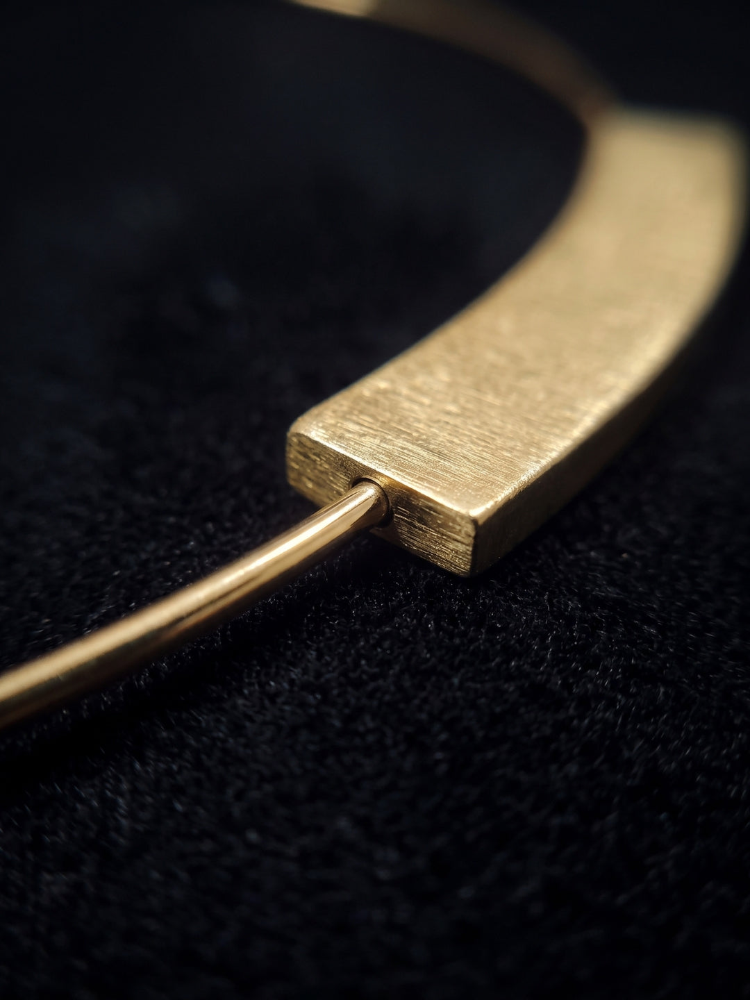 A close-up of a gold-toned metal necklace with a textured rectangular bar and a thin curved wire, resting on a black textured surface.
