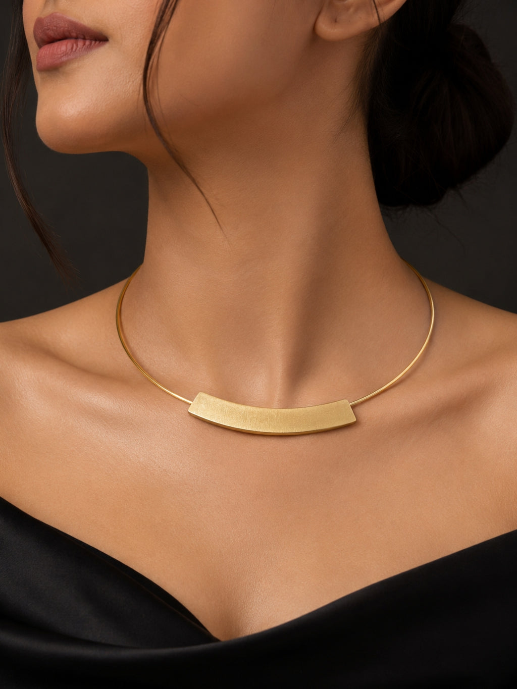 A close-up of a model wearing a gold collar necklace with a curved bar pendant, styled with a black off-the-shoulder top against a dark background.