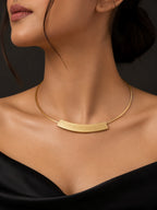 A close-up of a model wearing a gold collar necklace with a curved bar pendant, styled with a black off-the-shoulder top against a dark background.