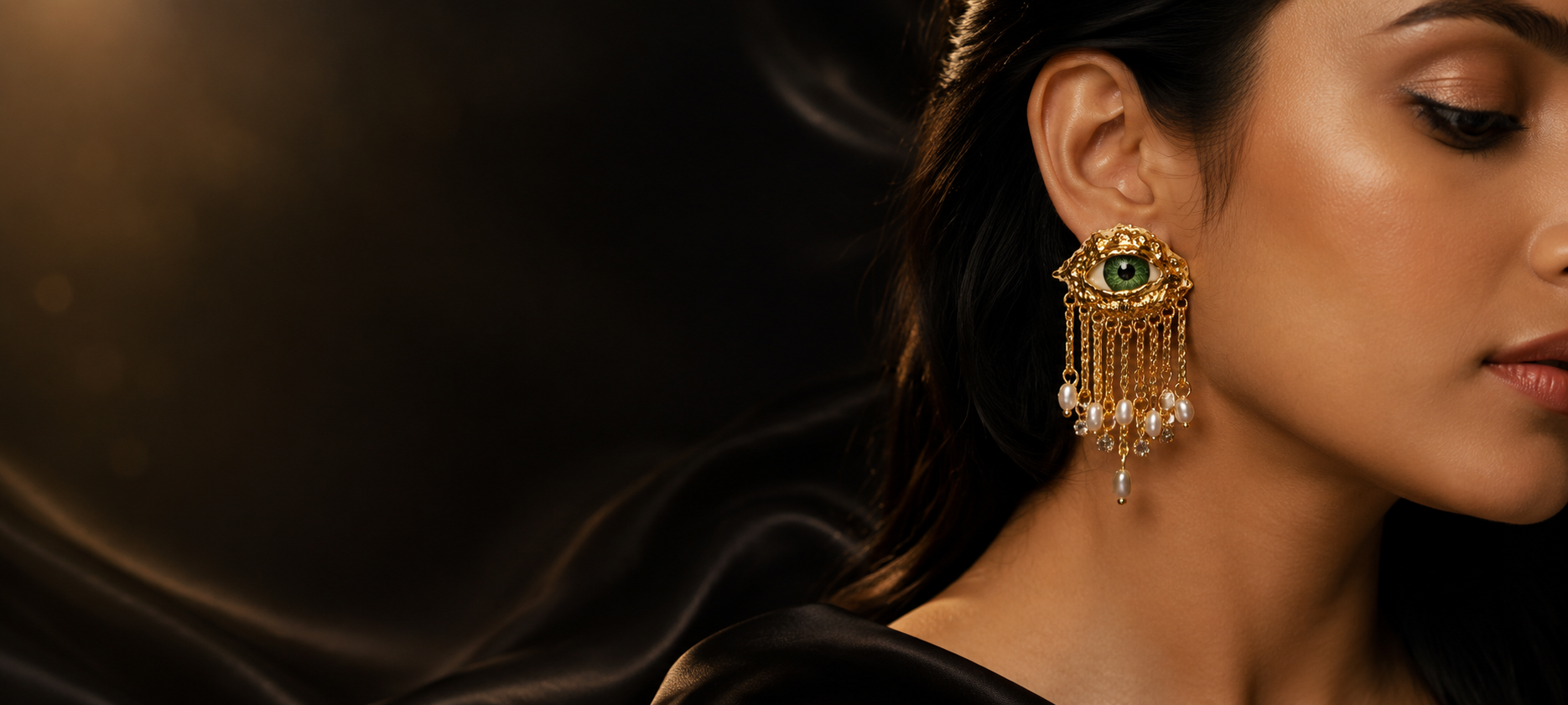 A close-up of a woman wearing a large ornate gold-toned earring with a green stone and dangling pearl-like accents, set against a dark draped background.
