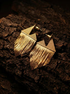 A pair of gold geometric dangle earrings with a textured triangular top and fringe design, displayed on a dark, rough bark surface.