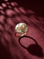 A gold ring featuring a large white pearl and diamond accents, displayed on a textured dark red surface with artistic leaf shadows.