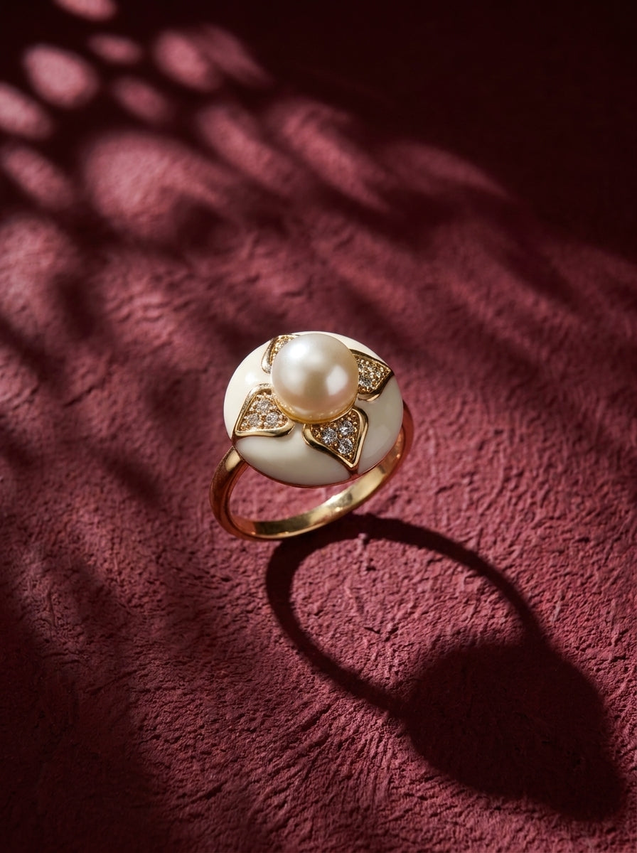 A gold ring featuring a large white pearl and diamond accents, displayed on a textured dark red surface with artistic leaf shadows.