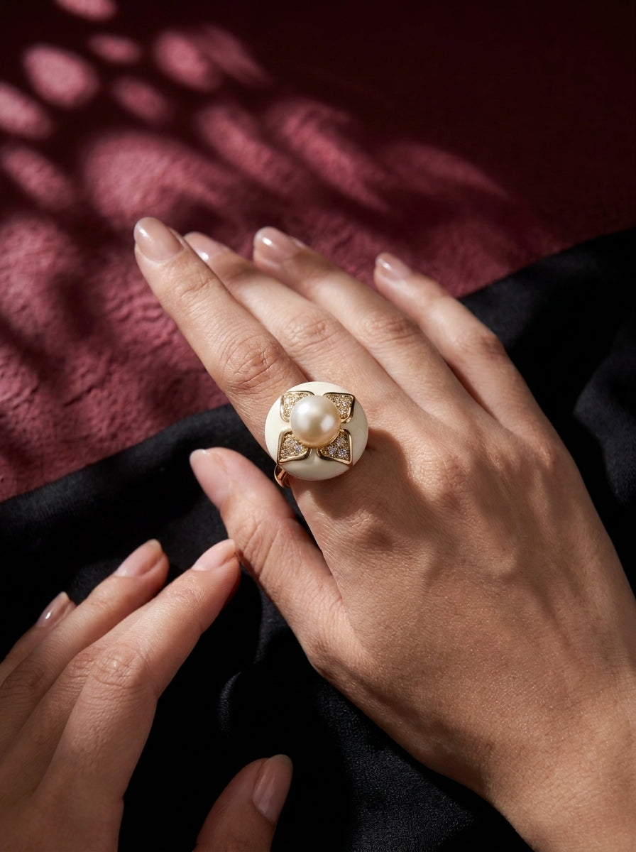 A close-up of a hand wearing a gold ring with a large white pearl and textured gold detailing, resting on black fabric with a dark red background showing soft shadow patterns.