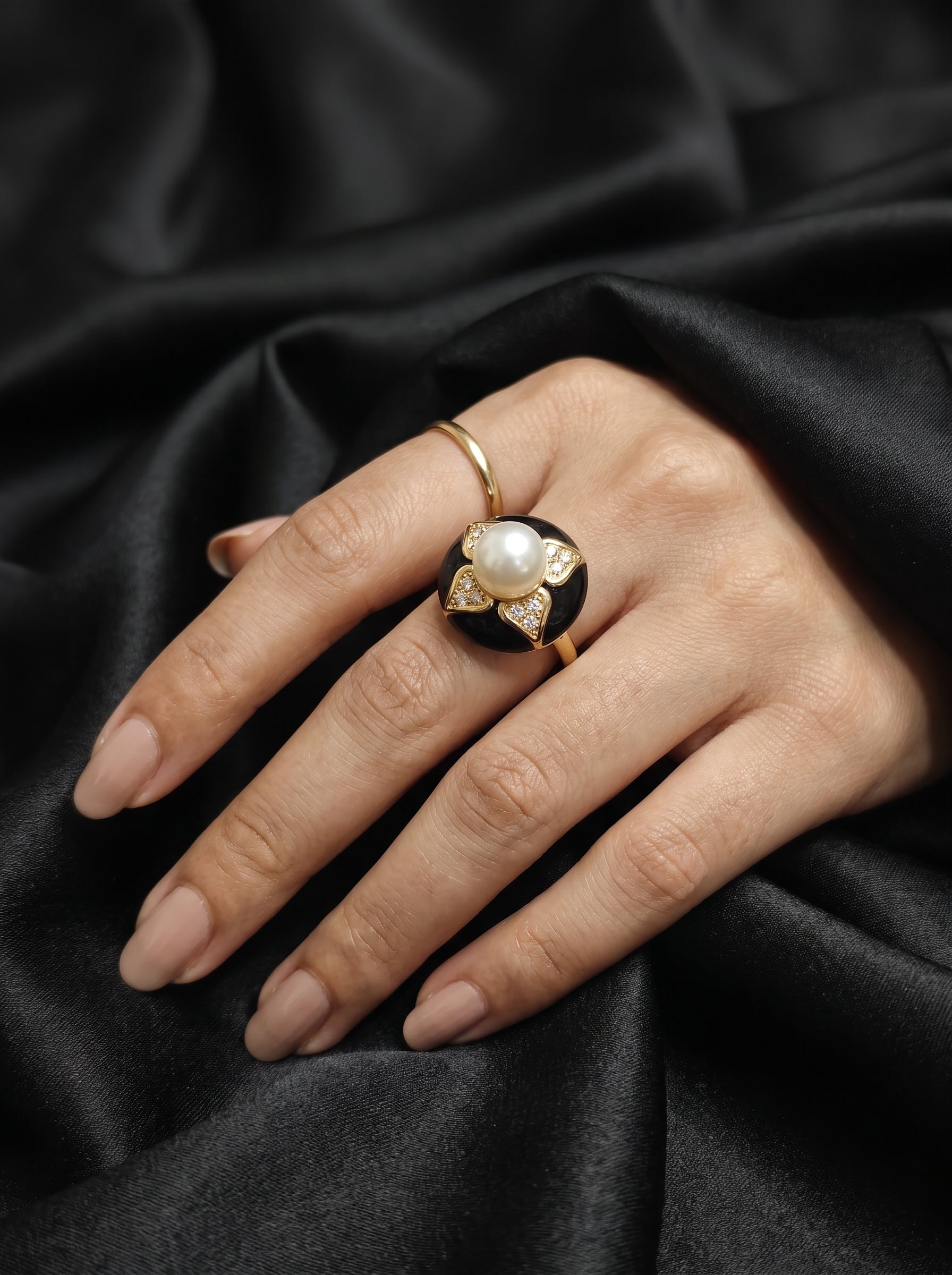 A close-up of a hand wearing a vintage-style gold ring with a large white pearl and black enamel details, resting on draped black satin fabric.