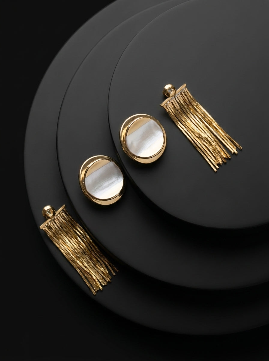 A pair of gold tassel drop earrings with oval white mother-of-pearl accents, displayed on a set of black circular pedestals against a dark background.