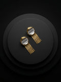A pair of gold-toned statement earrings with circular clear stone tops and long fringe, displayed on a dark round pedestal against a black background.
