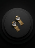 A pair of gold-toned statement earrings with circular clear stone tops and long fringe, displayed on a dark round pedestal against a black background.