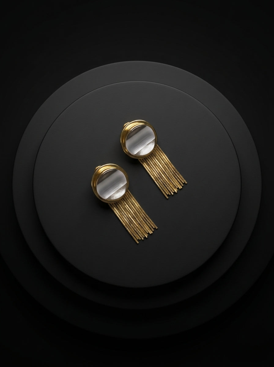 A pair of gold-toned statement earrings with circular clear stone tops and long fringe, displayed on a dark round pedestal against a black background.