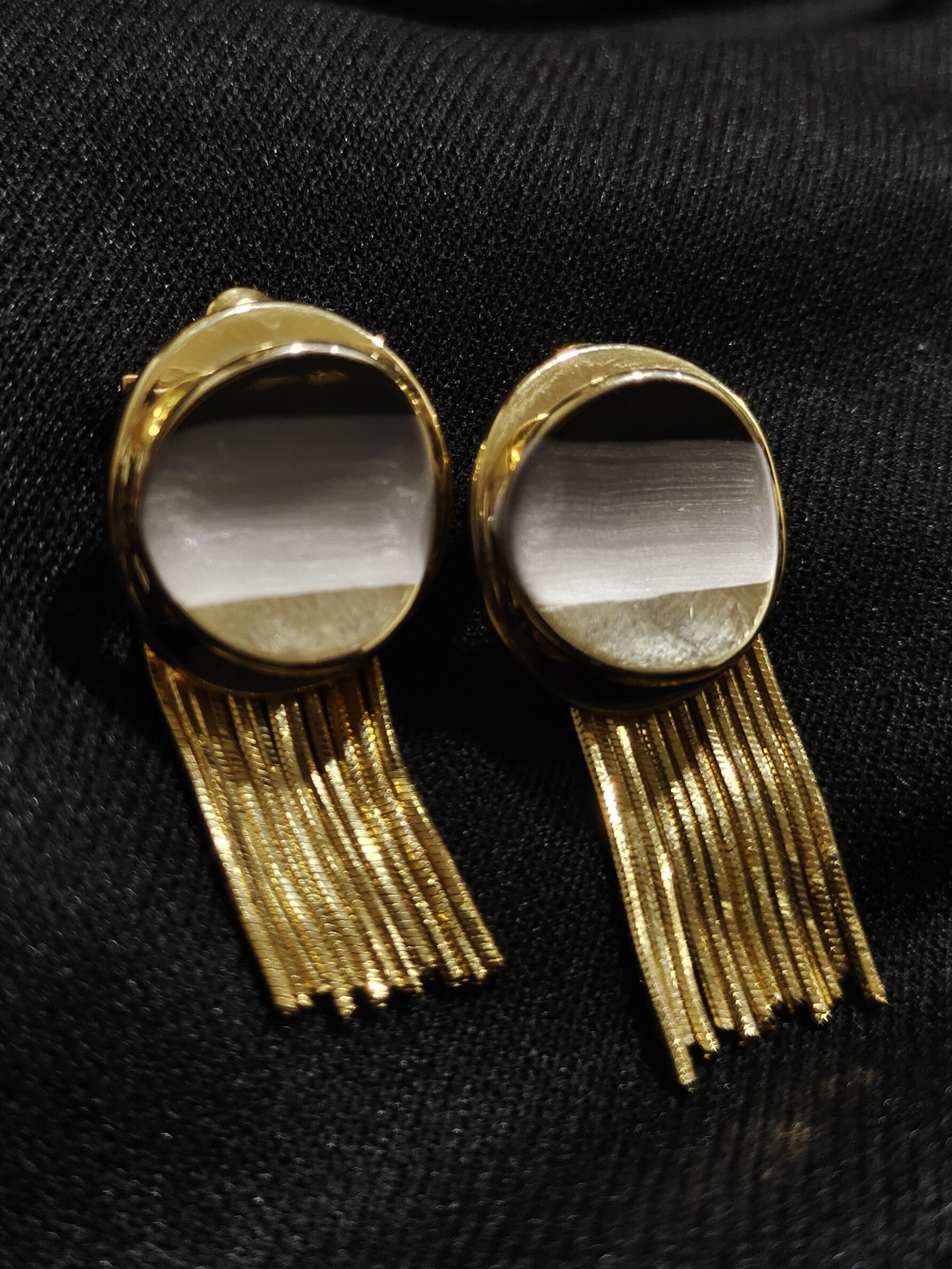 A pair of gold-toned statement earrings with oval cat-eye style stones and dangling fringe, displayed on a black textured fabric background.