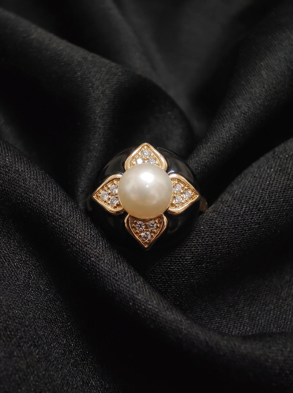 A gold ring featuring a central white pearl surrounded by four diamond-like accents, displayed on a draped black fabric background.