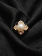 A gold ring featuring a central white pearl surrounded by four diamond-like accents, displayed on a draped black fabric background.