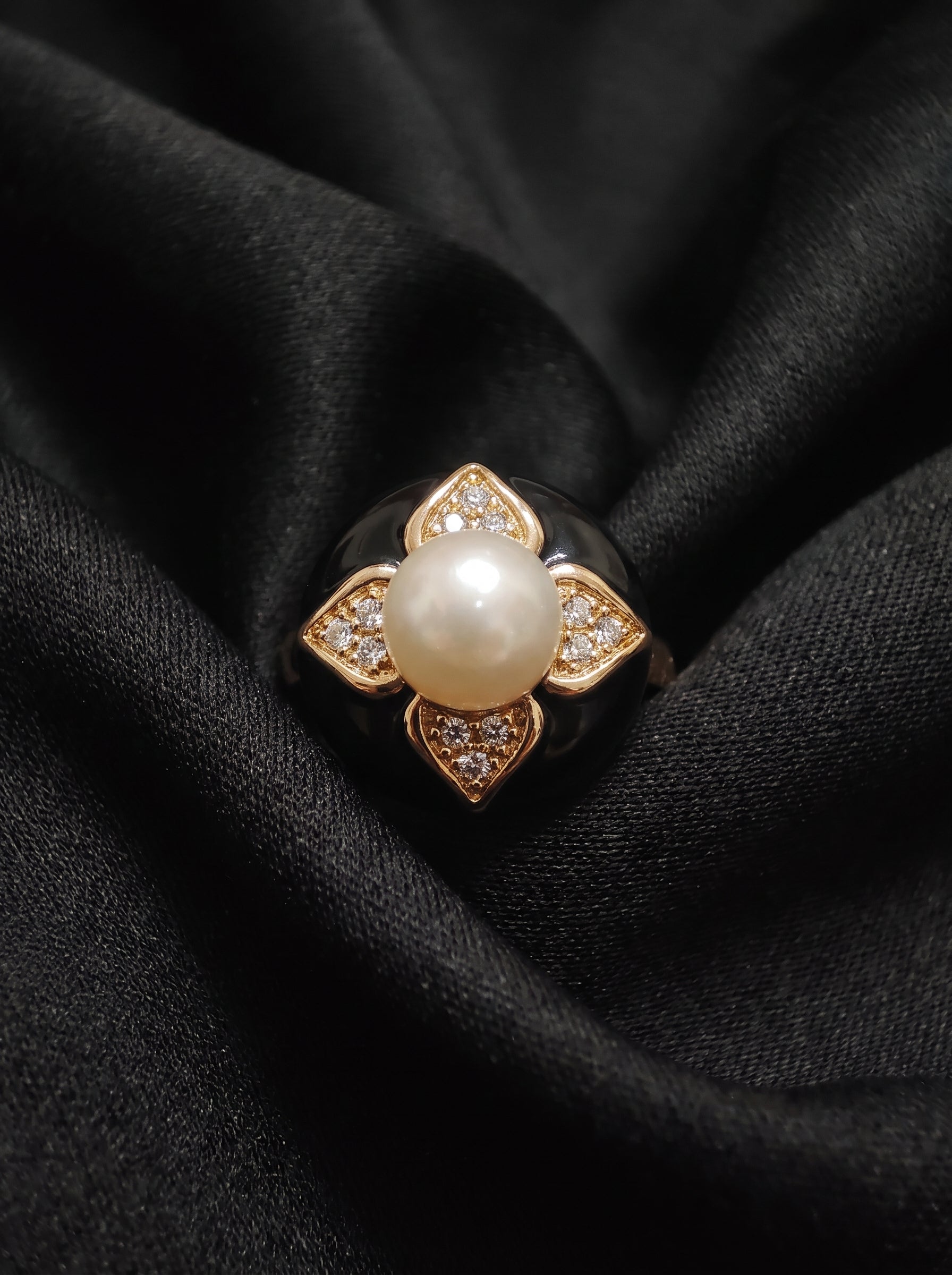 A gold ring featuring a central white pearl surrounded by four diamond-like accents, displayed on a draped black fabric background.