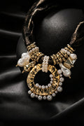 A statement necklace by Aadisha, featuring a large textured gold-tone circular pendant adorned with white pearls, accented with gold beads, a baroque pearl, and a faceted crystal, draped on a black satin background.
