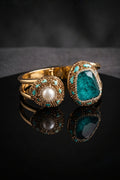 Two gold-plated statement rings displayed on a reflective black surface against a dark gray background. One ring features a round white pearl surrounded by small turquoise stones, and the other features a large, irregular-cut deep teal gemstone in a similar gold and turquoise halo setting.