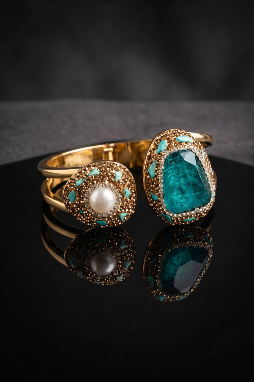 Two gold-plated statement rings displayed on a reflective black surface against a dark gray background. One ring features a round white pearl surrounded by small turquoise stones, and the other features a large, irregular-cut deep teal gemstone in a similar gold and turquoise halo setting.