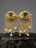A pair of ornate gold-toned earrings shaped like eyes with green iris details, dangling chains, and pearl-like drops, displayed on a dark stone surface against a gray studio background.