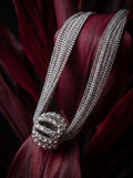 A close-up of a silver-toned multi-strand chain necklace with a round pavé-set centerpiece, resting on deep burgundy leaves against a dark background.