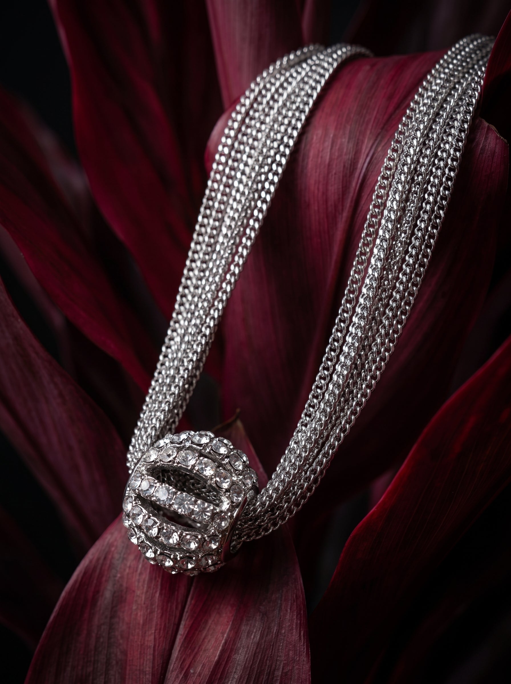 A close-up of a silver-toned multi-strand chain necklace with a round pavé-set centerpiece, resting on deep burgundy leaves against a dark background.